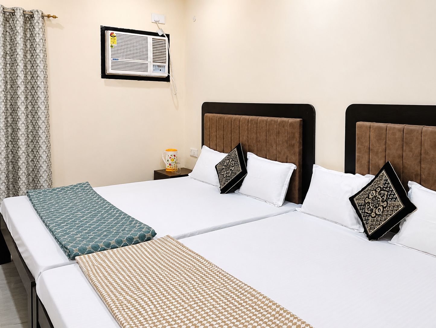 A hotel room with two beds, a nightstand with a lamp, a wall-mounted air conditioner, and a curtain.