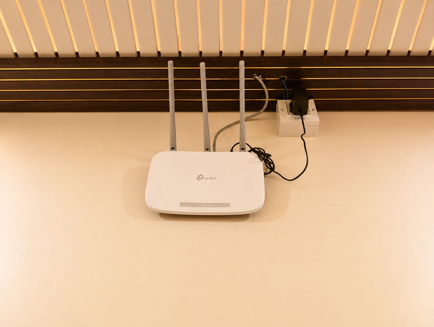 A white tp-link router with four antennas on a beige wall, connected to a power strip with a cable.