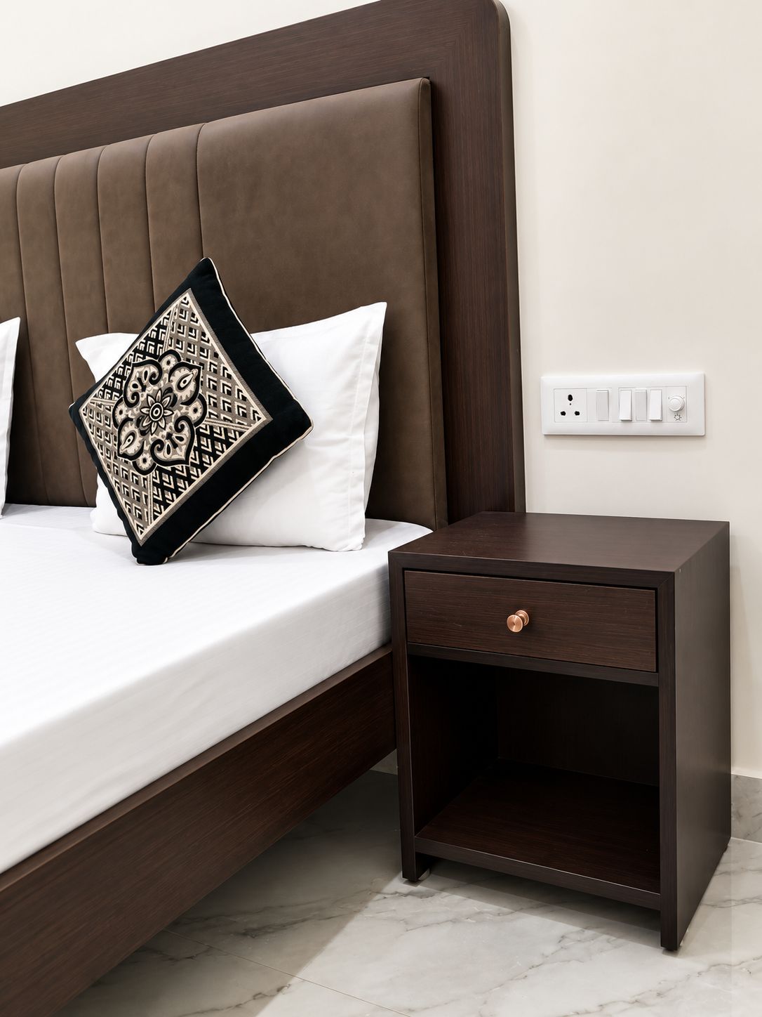 A bed with a headboard, a bedside table with a drawer, and a power outlet on the wall.