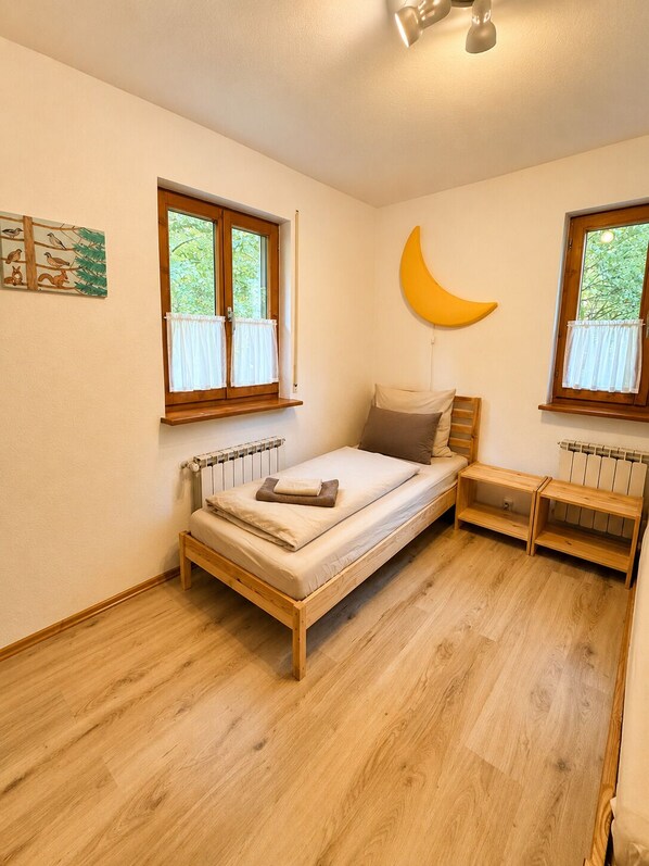 A small, single-bed room with a wooden frame, two windows with white curtains, a crescent moon decoration on the wall, and a radiator below one window.