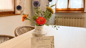 Une pièce avec une table dressée pour deux personnes, un vase rempli de fleurs, un panneau d’accueil, un four à micro-ondes et une fenêtre avec des rideaux.