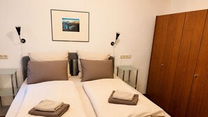 Un lit double avec deux oreillers et des serviettes pliées, une table de chevet, un tableau encadré au mur et une armoire.