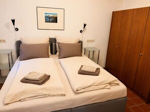 A double bed with two pillows and folded towels, a bedside table, a framed picture on the wall, and a wardrobe. - Löwenzahn Medical Practice, a former rural doctor's office in the Franconian Lake District—accommodates 6 people (Westheim)