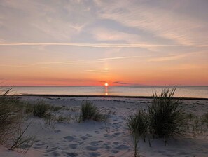 Ein Strand mit Sand und vereinzelten Grashalmen, ein ruhiges Meer und ein Sonnenuntergang.