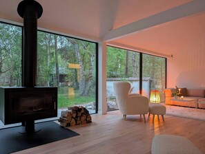 Ein modernes Wohnzimmer mit großem Kamin, einer gemütlichen Couch und Blick auf einen Wald durch bodentiefe Fenster.