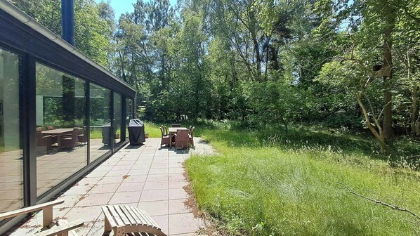 Une terrasse avec mobilier d’extérieur, un barbecue couvert et un espace clos par des parois vitrées, comprenant des tables et des chaises.