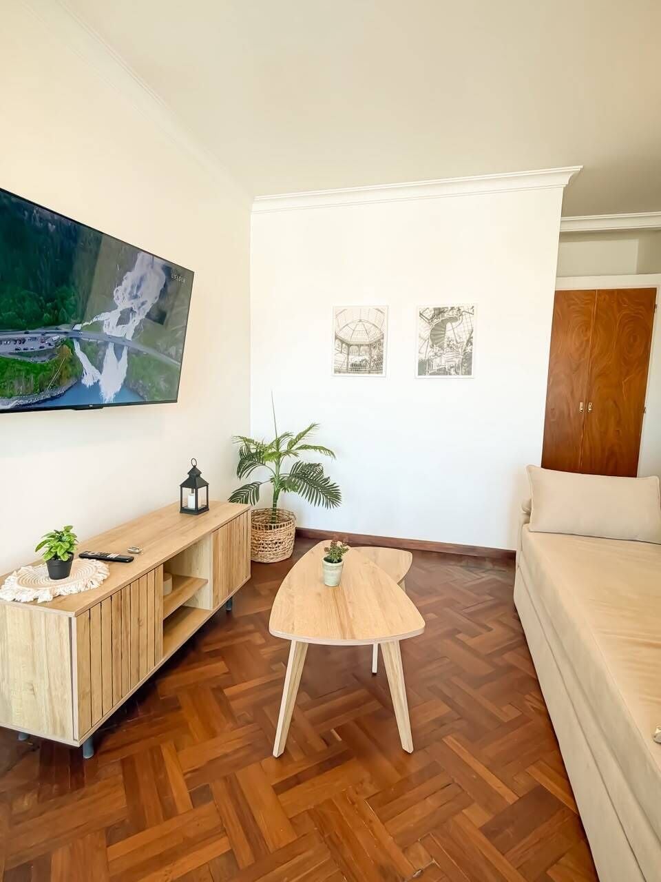 Ein Wohnzimmer mit einem großen Flachbildfernseher an der Wand, einer Holzunterhaltungsecke, einem runden Couchtisch, einem beigefarbenen Sofa und zwei gerahmten Bildern.