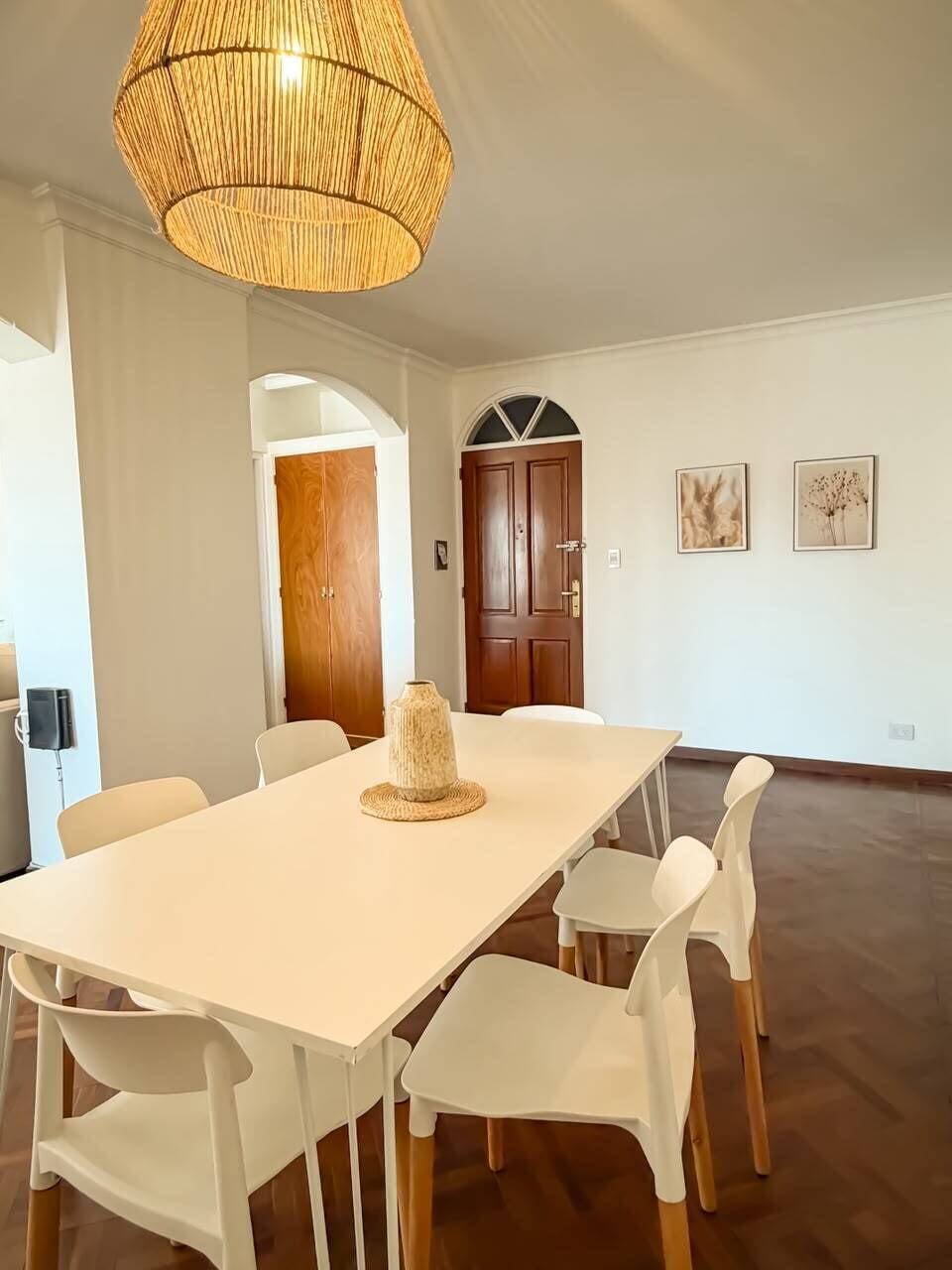 A dining area with a white table and chairs, a woven light fixture, and a wooden door in the background.