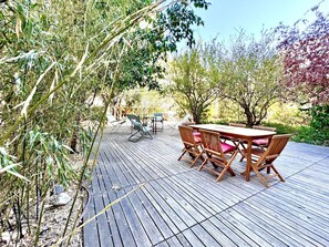 Une terrasse extérieure en bois, avec une table et des chaises, entourée de différentes plantes et arbres.