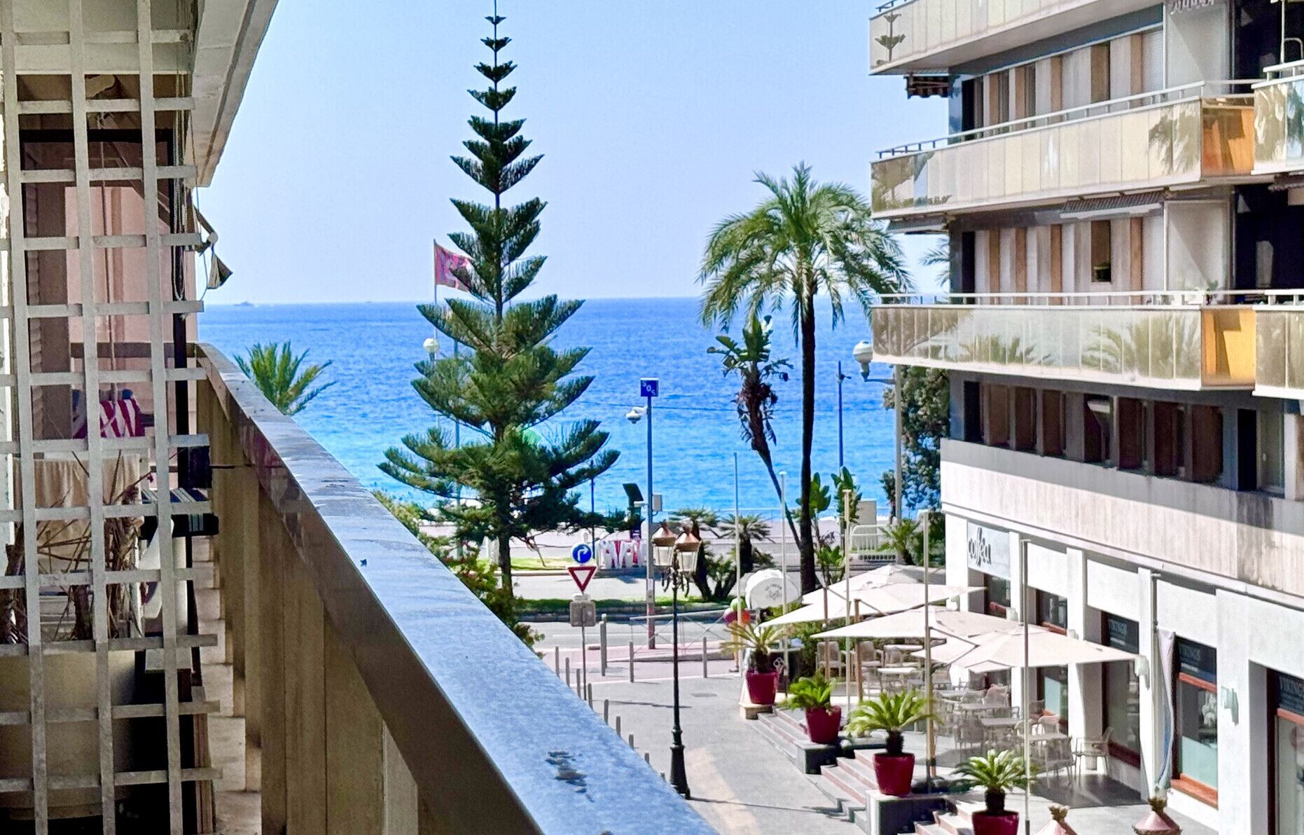 A coastal view from a balcony overlooking a street with palm trees and a clear blue sea.
