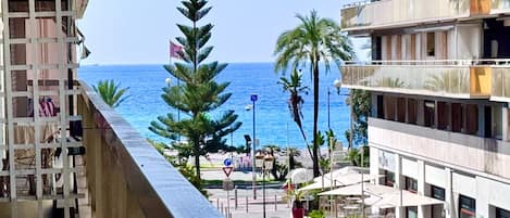 A coastal view from a balcony overlooking a street with palm trees and a clear blue sea.