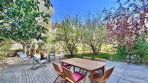 Eine Holzterrasse im Freien mit Tisch und Stühlen, umgeben von Bäumen und Grünpflanzen.