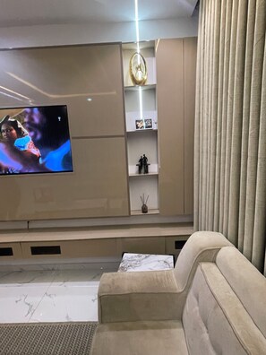 Ein Wohnzimmer mit flachbildfernseher an der Wand, einem beigefarbenen Sofa und einer dekorativen Leuchte.