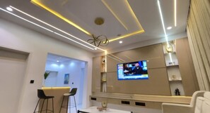 Ein modernes Wohnzimmer mit einem Wand-Flachbildfernseher, einer Deckenleuchte in Linienform und einer Sitzecke mit zwei Sesseln.