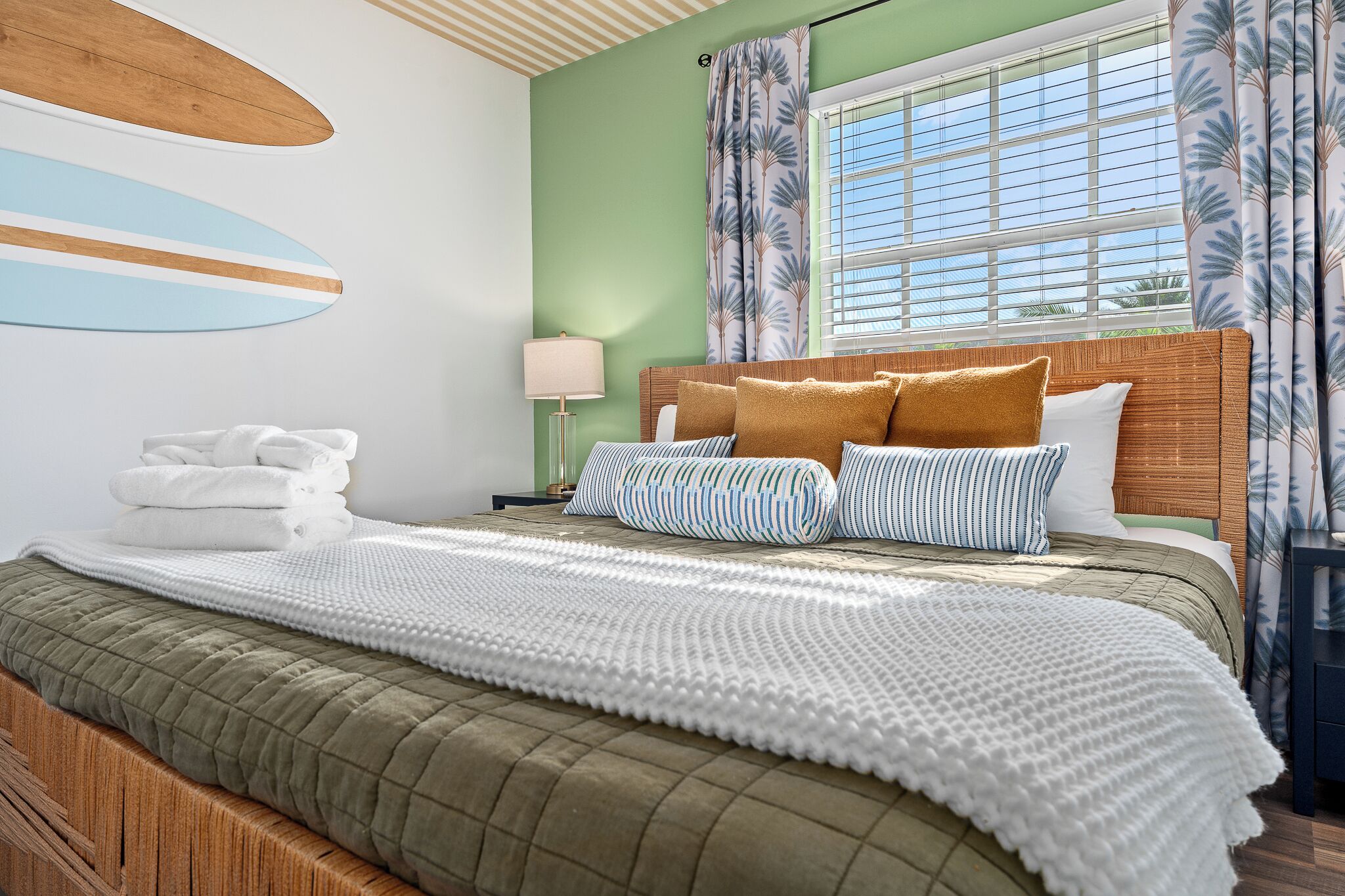 A hotel room with a bed, pillows, a blanket, a nightstand, a lamp, and a window with curtains.