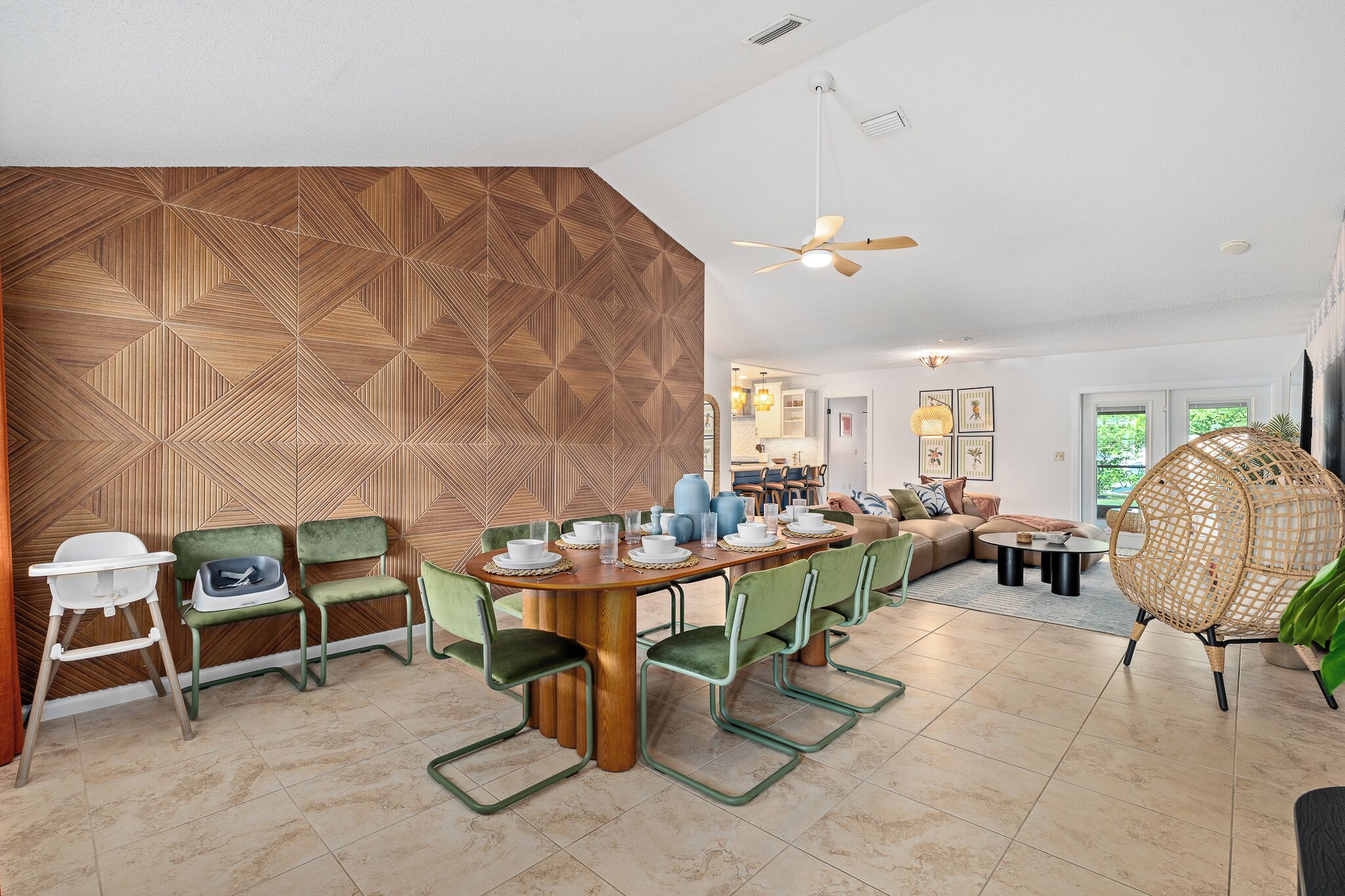 A modern dining area with a geometric patterned wall, green upholstered chairs, a wooden dining table, and a ceiling fan.