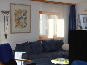 Une pièce avec un canapé gris, un fauteuil bleu, une petite table ronde avec une lampe et un tableau accroché au mur.