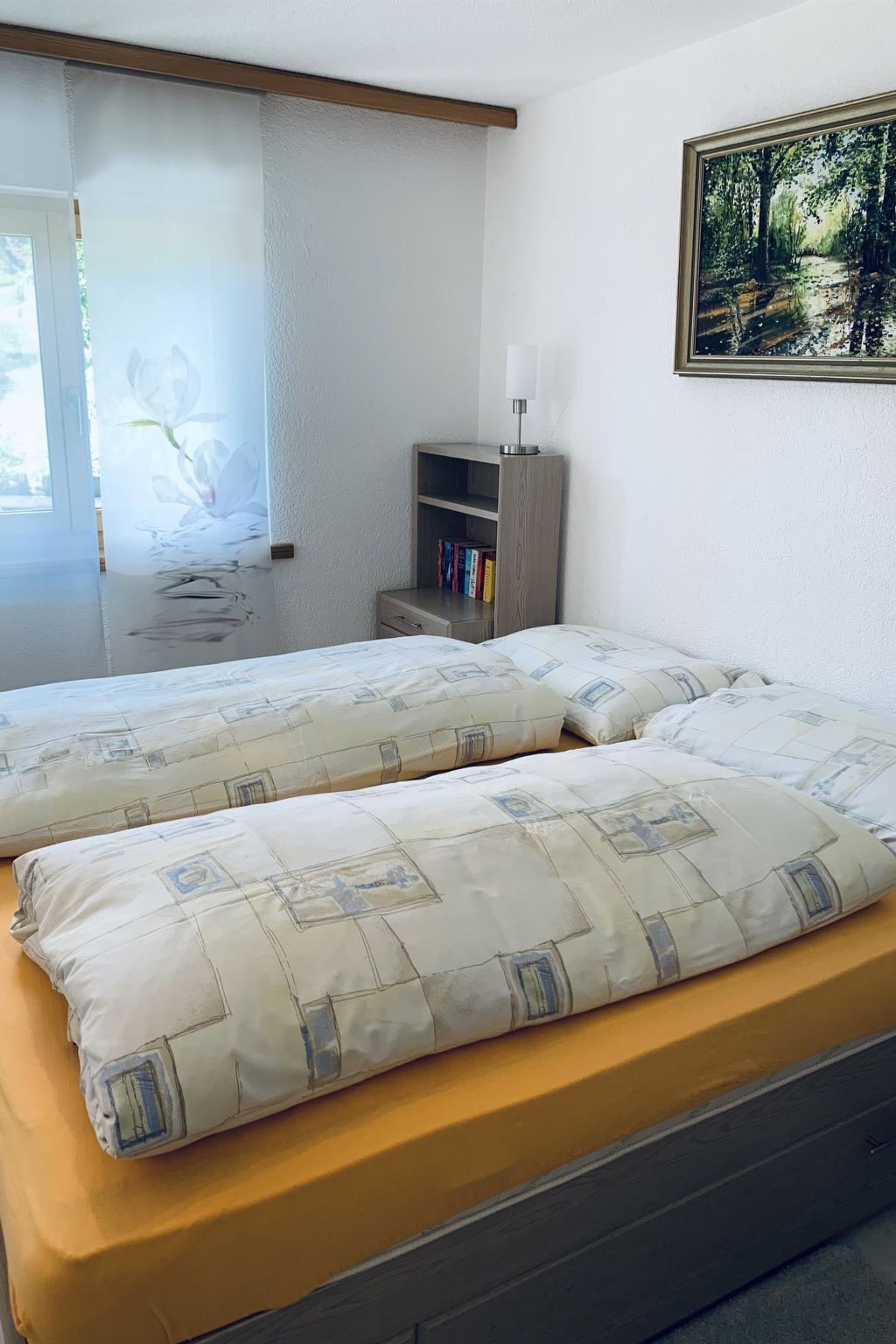 Quarto com duas camas, uma mesa de cabeceira com livros, uma luminária e um quadro na parede.