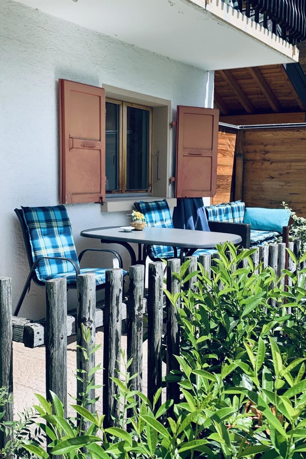 En utomhusplats med ett bord och stolar, ett fönster med fönsterluckor och ett trästaket.