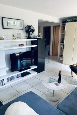 Un salon équipé d’un téléviseur à écran plat, d’un ventilateur, d’une table basse avec une bouteille et d’une bibliothèque.