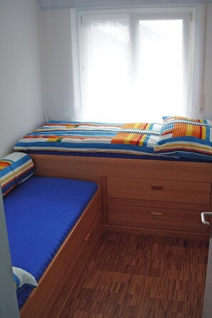 Ein kleines Zimmer mit einem Einzelbett, Holzboden, einem Fenster mit Vorhängen und einer farbenfrohen, gestreiften Bettdecke.