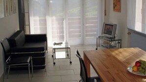 Ein Zimmer mit einer schwarzen Ledersofa, einem Holzesstisch mit einer Fruchtschale, einem Fernseher auf einem Glastisch und Blick durch große Fenster.