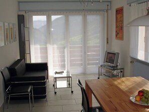 Ein Zimmer mit einer schwarzen Ledersofa, einem Holzesstisch mit einer Fruchtschale, einem Fernseher auf einem Glastisch und Blick durch große Fenster.