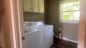 A laundry room with a washer and dryer, wooden flooring, a window with blinds, and a door leading to another room.