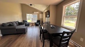 A spacious living room with a dark-colored sofa, a dining table with chairs, and a large window providing a view of the outdoors.