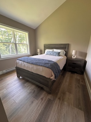 A bedroom with a large window, a wooden bed, two bedside tables with lamps, and a wooden floor.