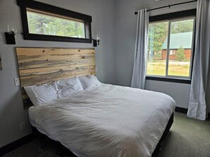 Une chambre à coucher avec une tête de lit en bois, un lit avec une literie blanche, une fenêtre avec des rideaux et une vue sur l’extérieur.