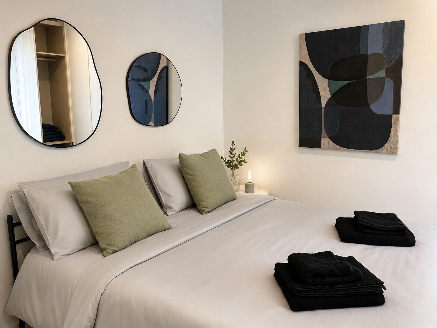 A neatly made bed with pillows and a folded towel, a bedside table with a lamp, and two wall mirrors.
