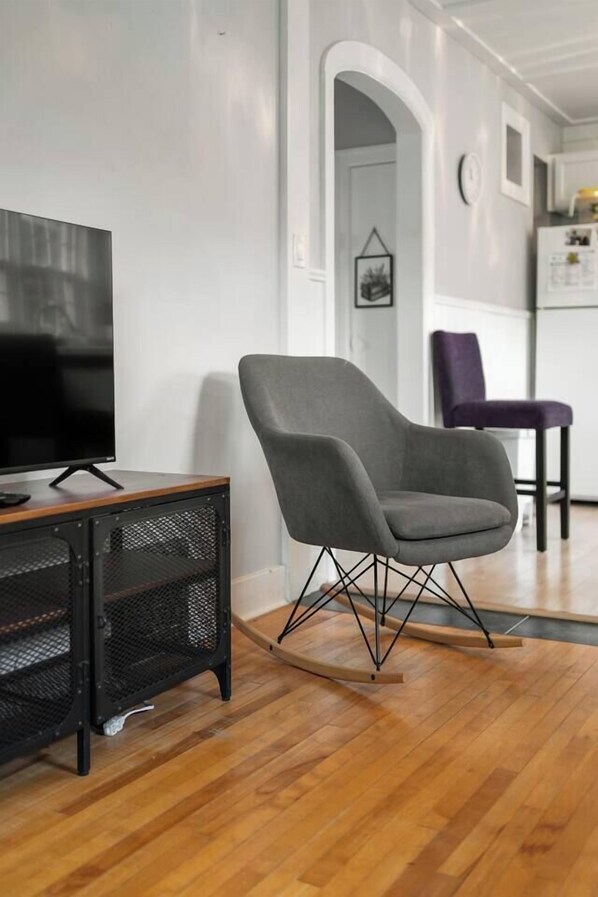 A modern living room with a flat-screen TV on a wooden stand, a grey upholstered chair, and a black mesh speaker.