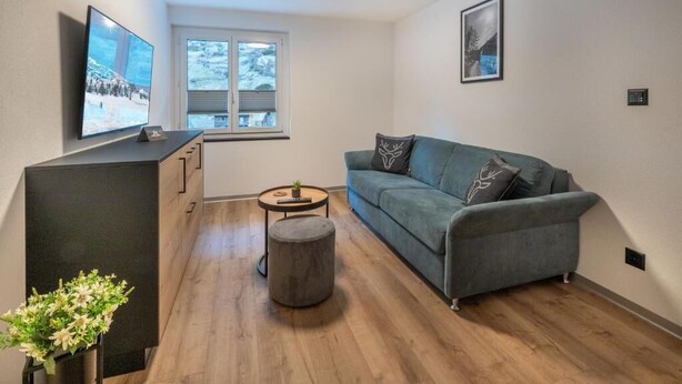 Ein modernes Wohnzimmer mit grauem Sofa, Flachbildfernseher, kleinem runden Tisch und einem Fenster mit Jalousien.