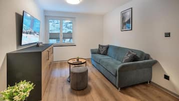 Ein modernes Wohnzimmer mit grauem Sofa, Flachbildfernseher, kleinem runden Tisch und einem Fenster mit Jalousien.
