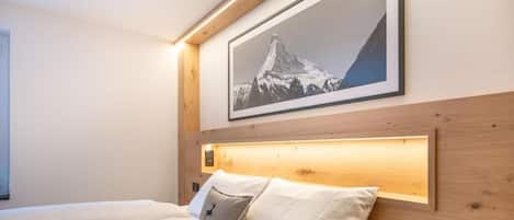 A hotel room with a bed, a headboard with a built-in light, and a framed picture of a mountain on the wall.