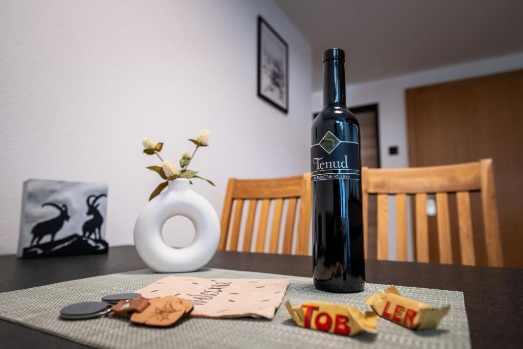 Una botella de vino Tenud, un jarrón blanco con flores secas, un cuadro enmarcado y una silla de madera.