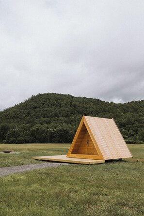 Una estructura triangular de madera en un campo cubierto de hierba, con una colina boscosa al fondo.