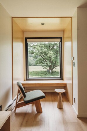 Una habitación con un banco empotrado y una mesita pequeña, un ventanal con vistas a árboles y suelo de madera.