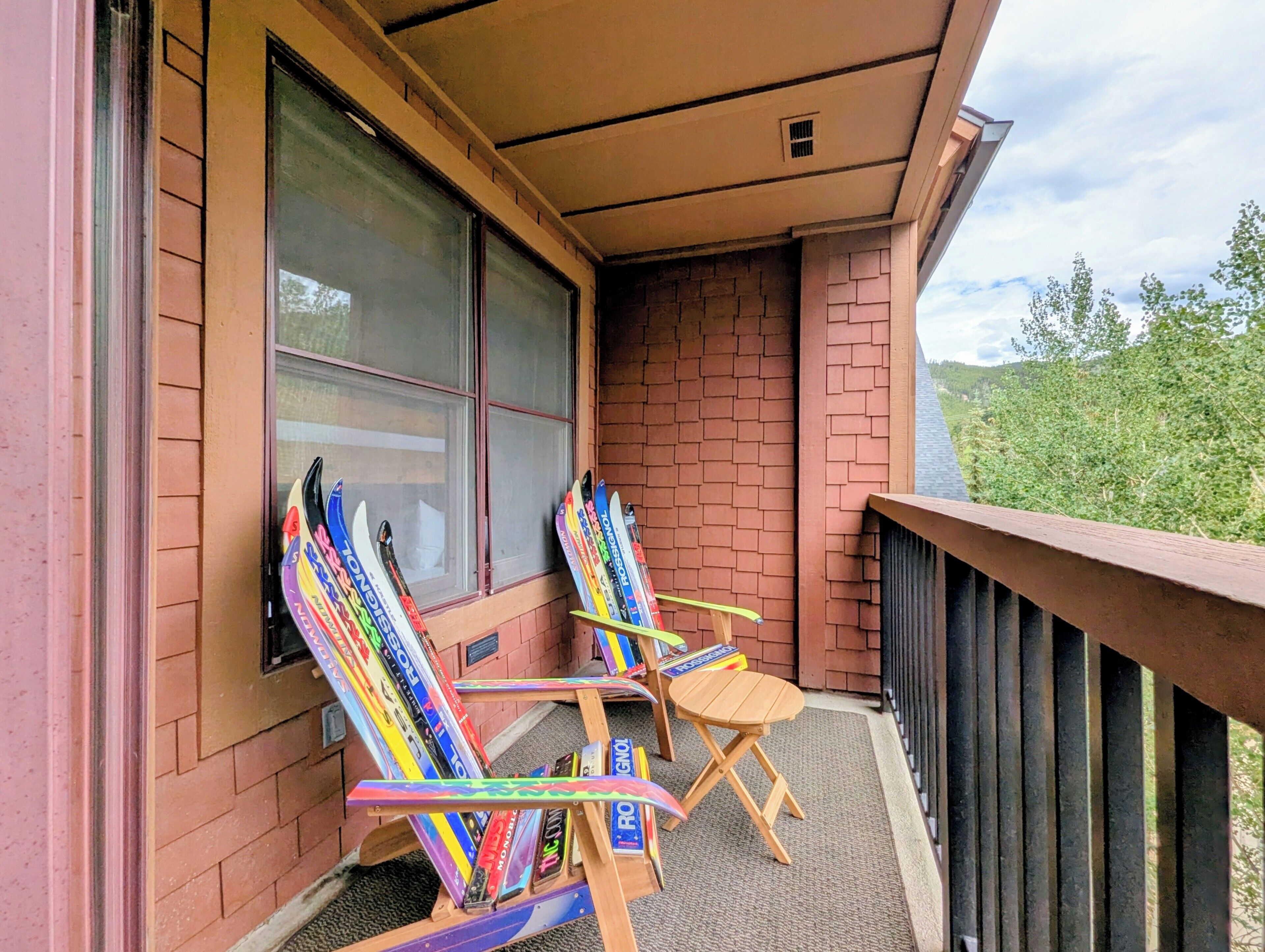 A balcony with two wooden chairs and a table, a window with blinds, and a brick wall.