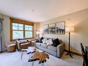 A living room with a sofa, two armchairs, a coffee table, and a floor lamp. There are three wall-mounted paintings, a window with blinds, and a fireplace. - New Listing | King Bed | Steps from the slopes (Keystone)