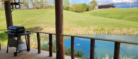 A wooden deck with a grill and a gas tank, overlooking a grassy field and a body of water.