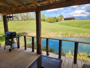 A wooden deck with a grill and a gas tank, overlooking a grassy field and a body of water. - Relaxing 1-bedroom cabin overlooking duck pond in charming West Liberty (WEST LIBERTY)