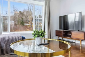 A modern living room with a large window, a round glass coffee table, a television, and a wooden cabinet.