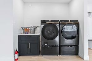 A laundry room with two front-loading washers and a dryer, a small cabinet, a basket with detergent, and a fire extinguisher.