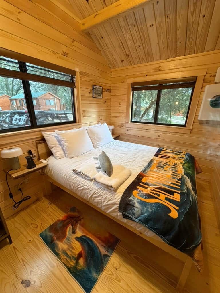 Quarto de cabana de madeira com cama, janelas, mesa de cabeceira e um quadro no chão.