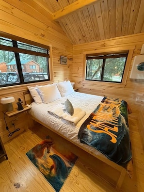 A wooden cabin bedroom with a bed, windows, a nightstand, and a painting on the floor.