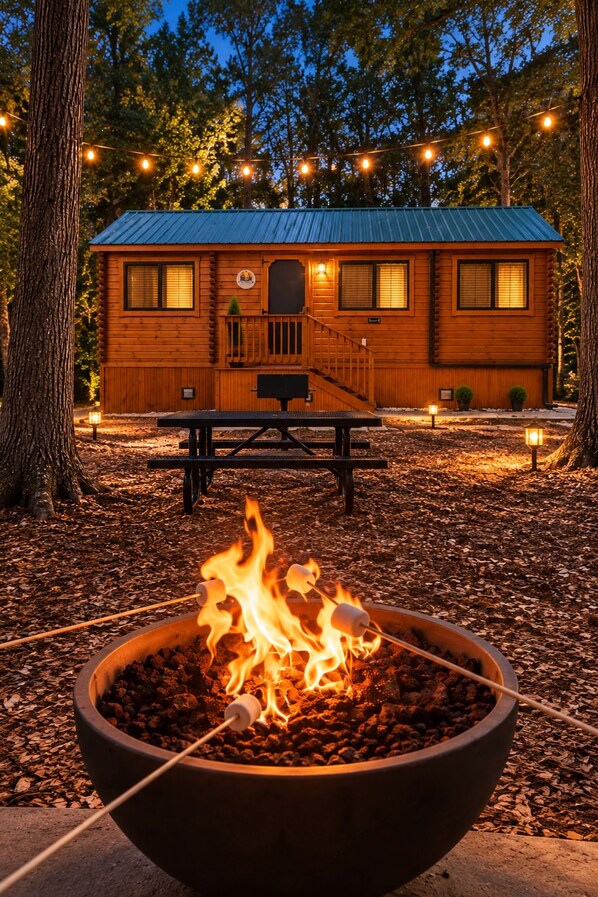 A rustic cabin with a fire pit in the foreground and string lights in the background.
