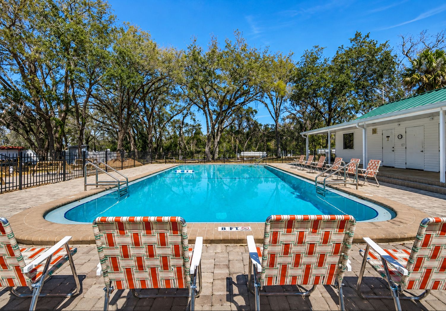 A rectangular swimming pool with a tiled deck, surrounded by trees and a fenced area. There are lounge chairs and a covered seating area with a green roof.