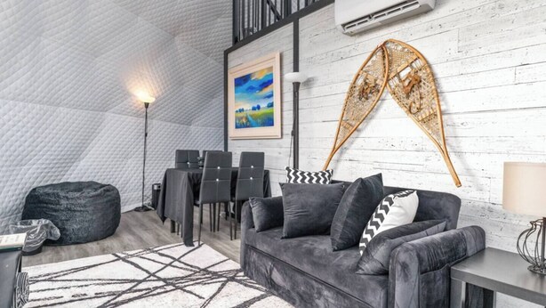 Ein Innenraum mit hoher Decke, einer Essecke mit Stühlen und Tisch, einer Couch mit Kissen, einer Lampe und einem Gemälde an der Wand.